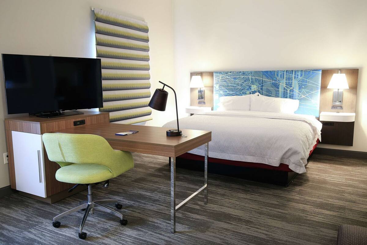A hotel room with a bed, a desk with a chair, a television, two bedside lamps, and a large window with a view.