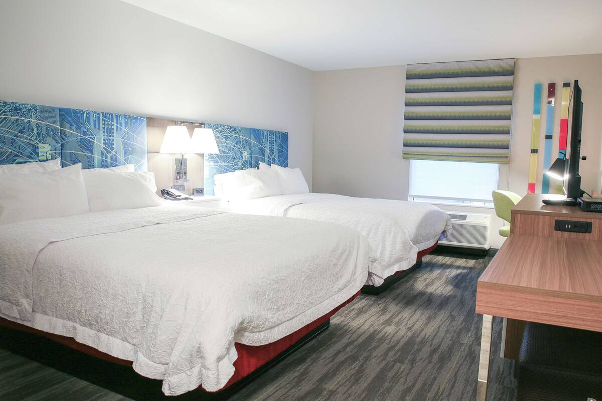 A hotel room with two beds, a desk, a lamp, and a large window with blinds.