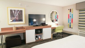 A hotel room with a bed, a desk with a flat-screen TV, a green chair, a small table, a microwave, and a framed picture on the wall.