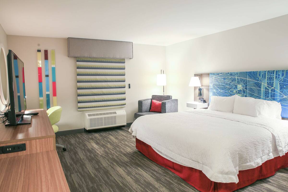 A hotel room with a large bed, a desk with a computer, a chair, a wall-mounted air conditioner, and a decorative wall panel.