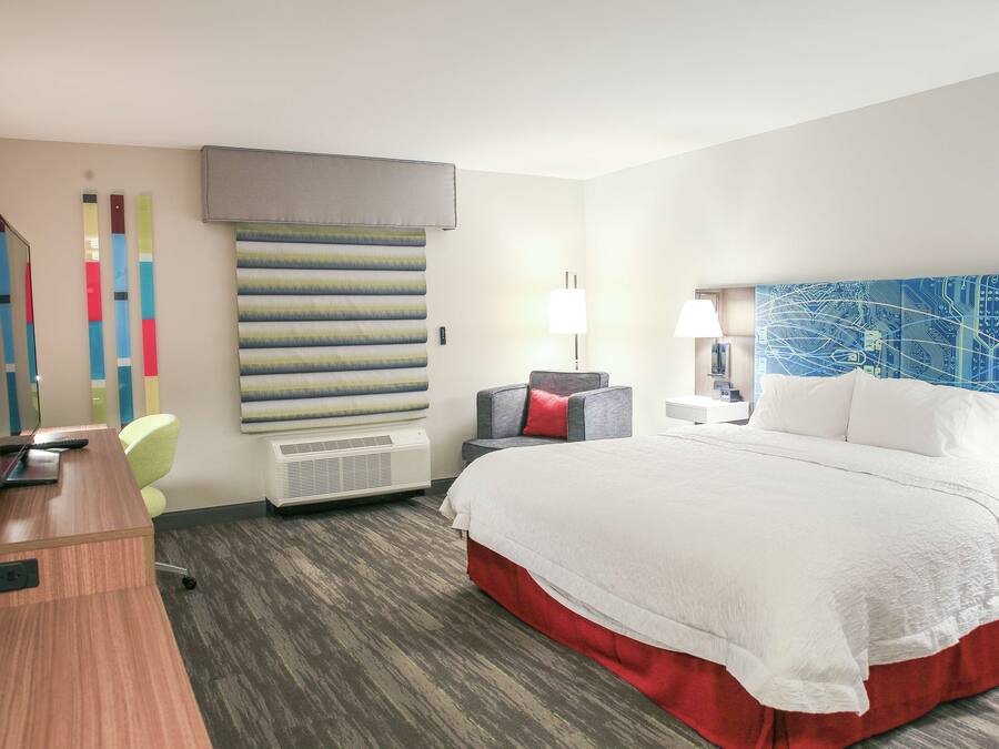 A hotel room with a large bed, a desk with a computer, a chair, a wall-mounted air conditioner, and a decorative wall panel.