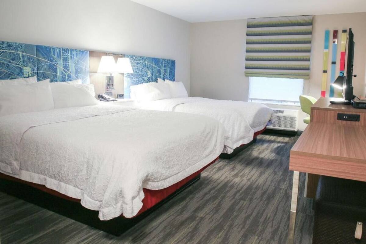 A hotel room with two beds, a desk, a lamp, and a large window with blinds.
