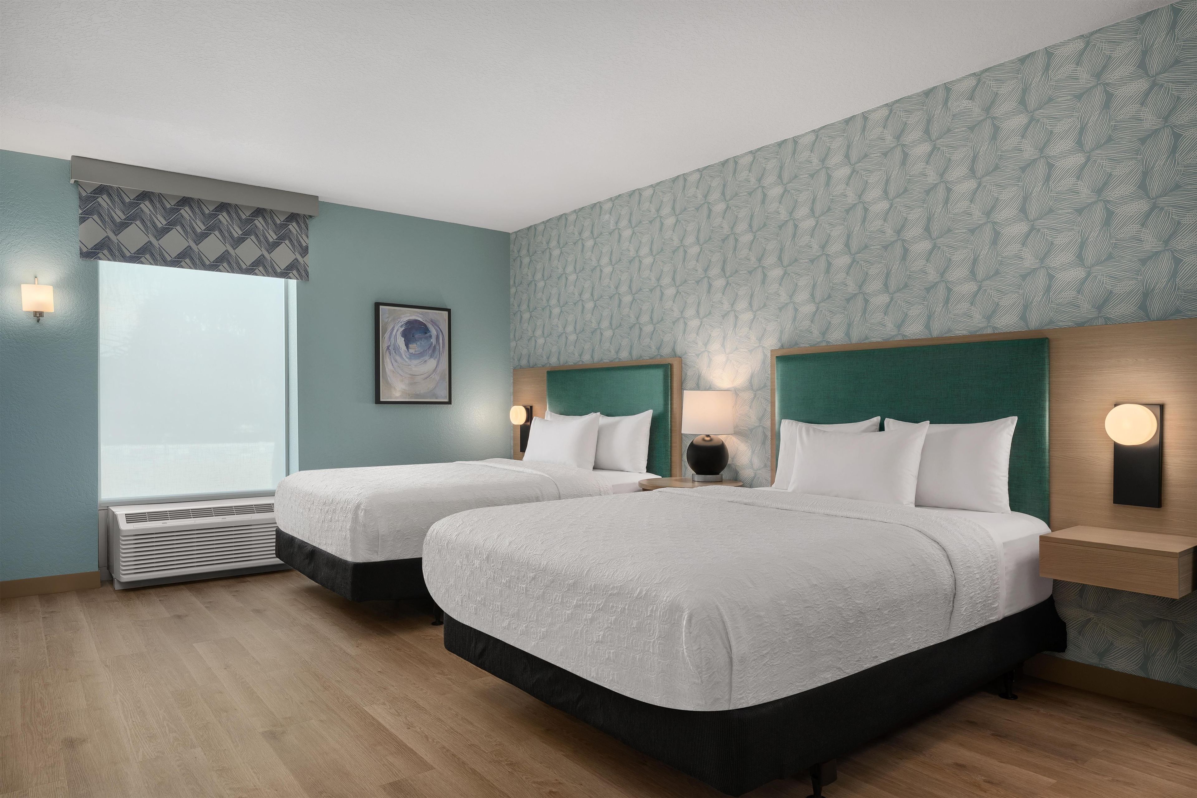 A hotel room with two beds, a large window, a wall-mounted air conditioner, and a painting.