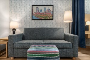 A room with a grey sofa, a patterned ottoman, a framed cityscape picture, two floor lamps, and a patterned wallpaper.