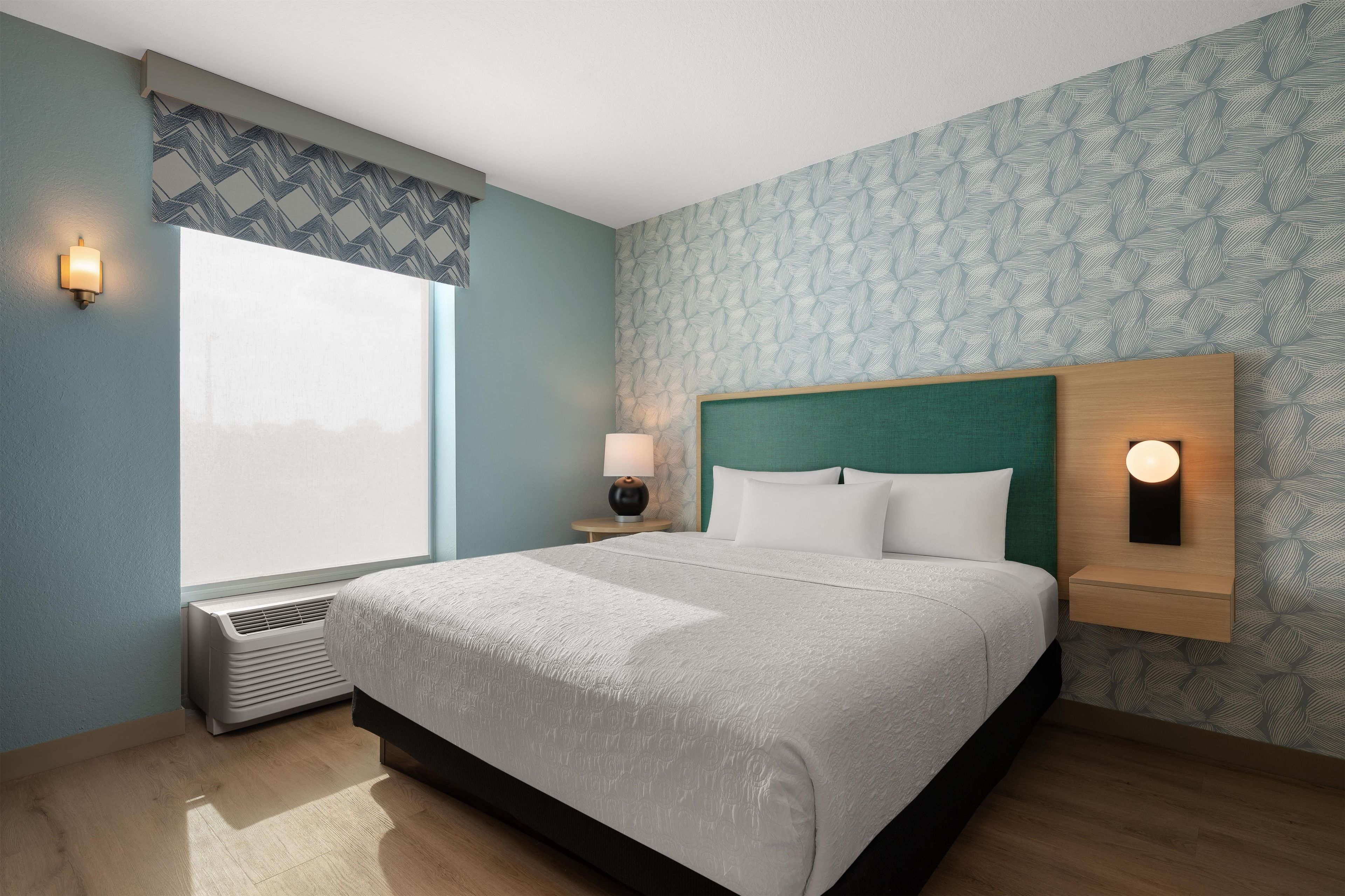 A hotel room with a large bed, a nightstand with a lamp, a wall-mounted light, and a window with a patterned curtain.