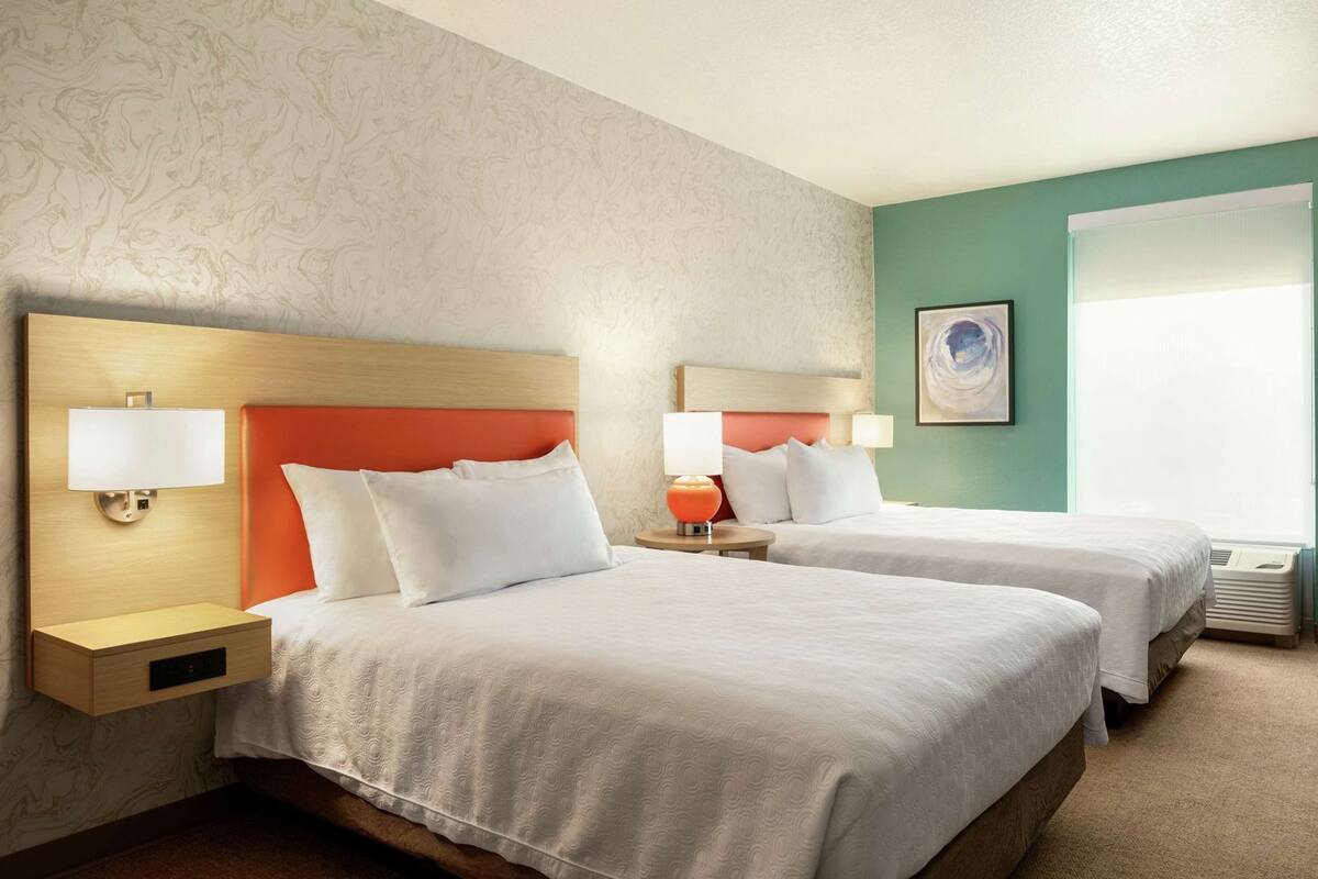 Een hotelkamer met twee bedden, nachtkastjes met lampen, een wandlamp en een raam met zonwering.