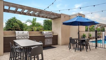 An outdoor patio with a barbecue grill, table, chairs, and a pool in the background.