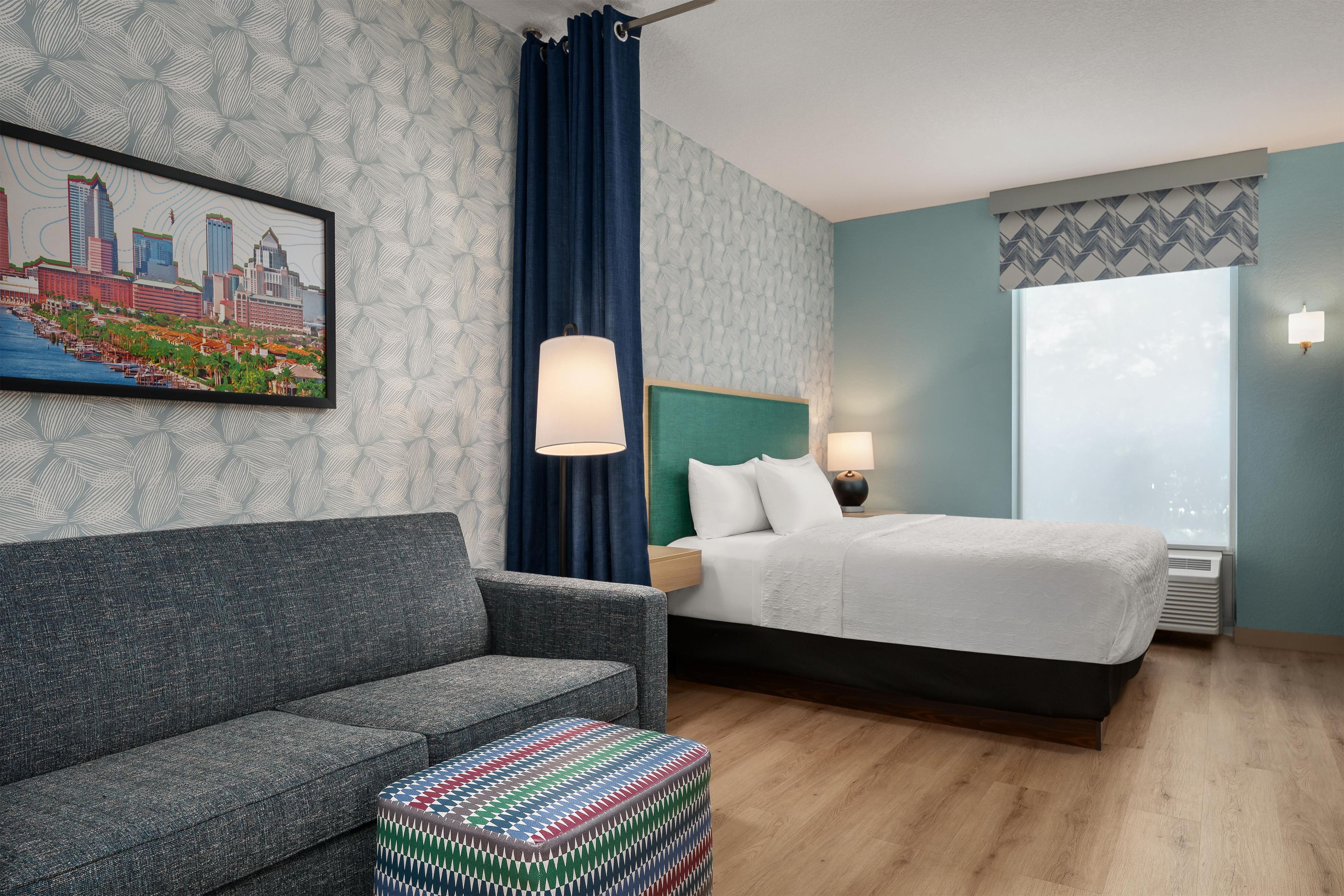 A hotel room with a bed, a sofa, a patterned ottoman, a cityscape painting, a window with curtains, and a wall with a leaf pattern.