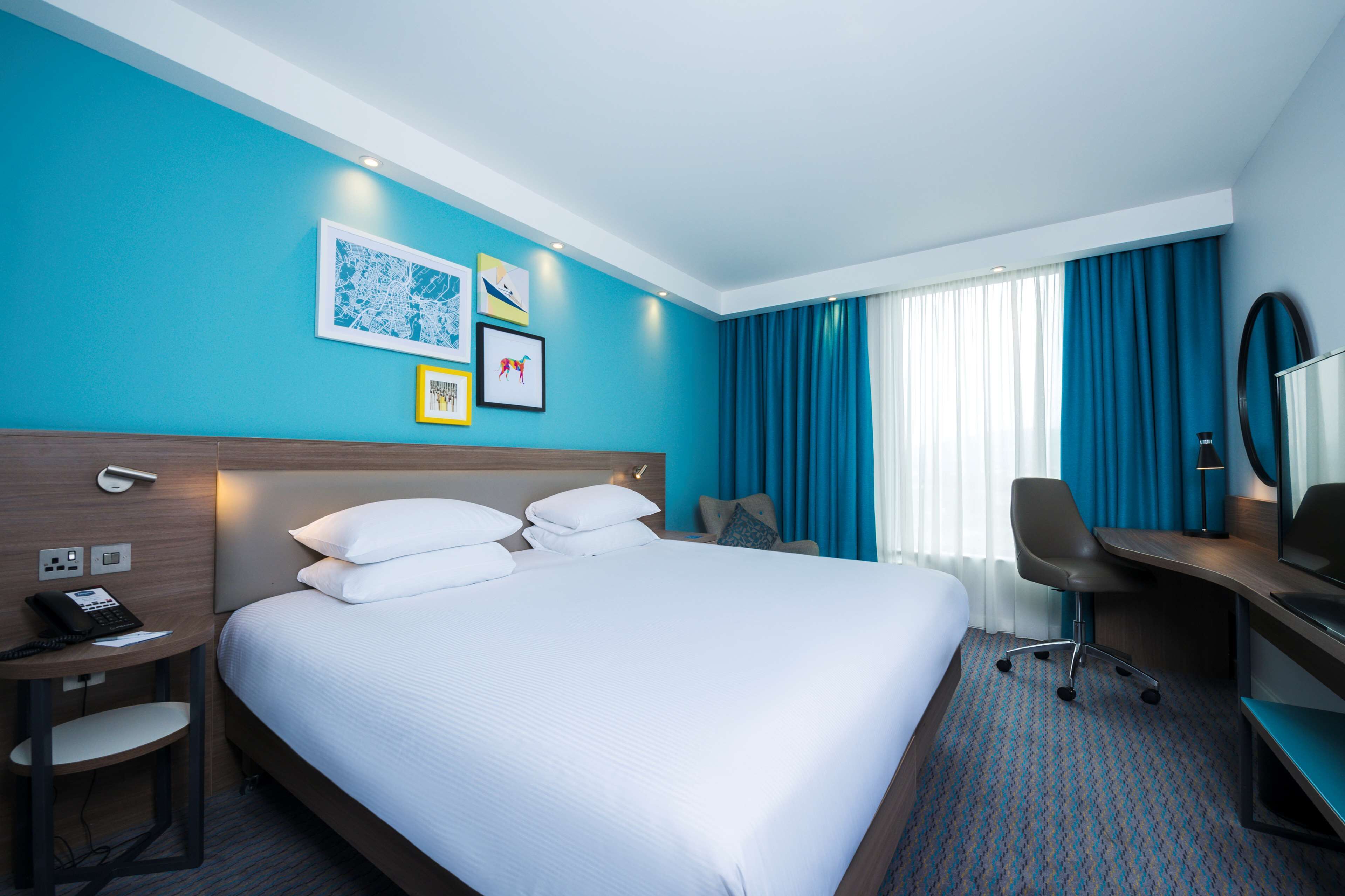 A hotel room with a large bed, a desk, a chair, and a desk lamp. There are framed pictures on the wall and a phone on the desk.