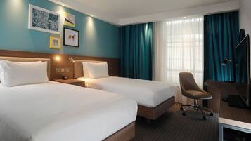 Quarto de hotel com duas camas, uma escrivaninha com cadeira, televisão e uma janela grande com cortinas.