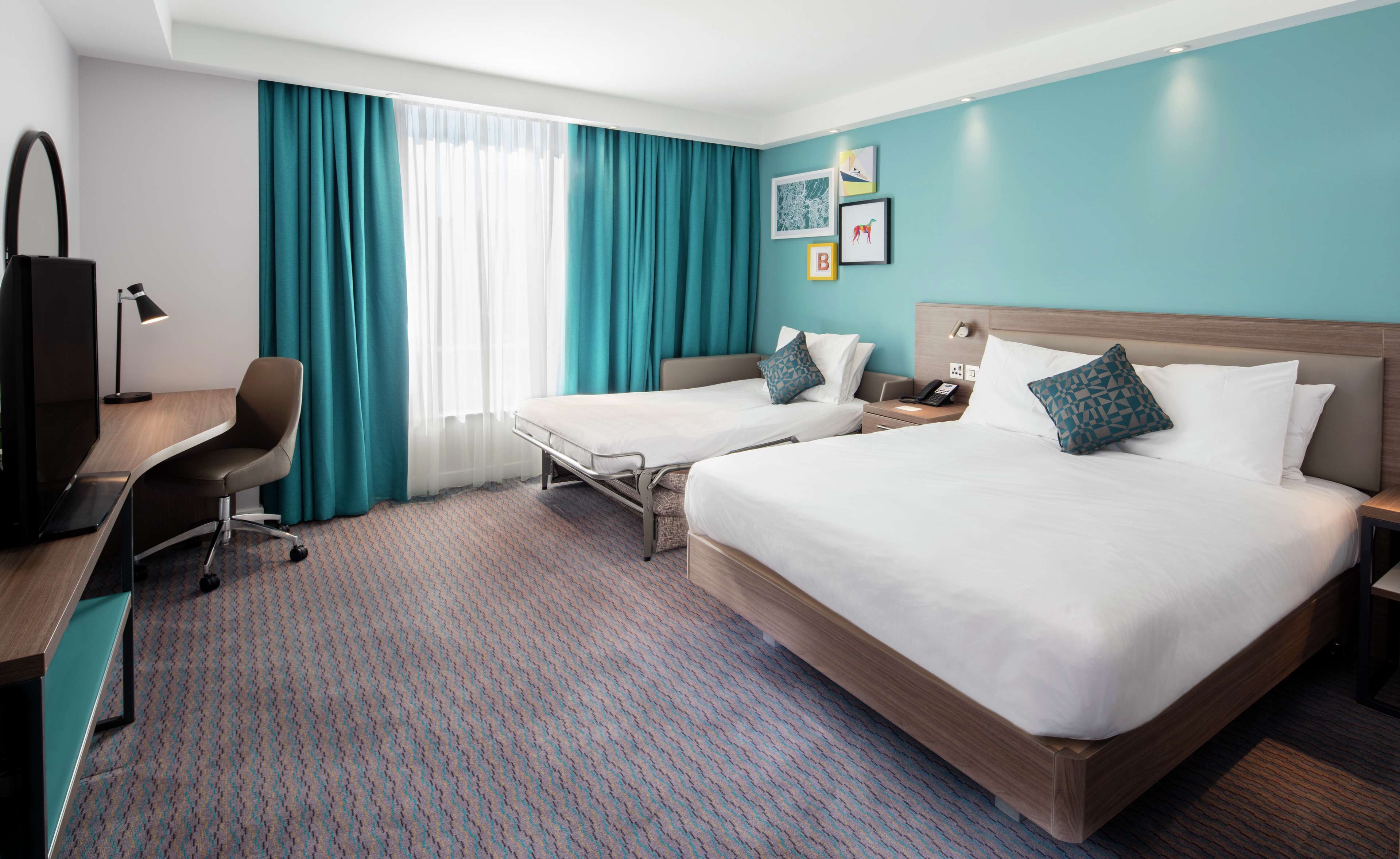 Ein Hotelzimmer mit zwei Betten, einem Schreibtisch, einem Stuhl, einer Schreibtischlampe und einem Fenster mit Vorhängen.