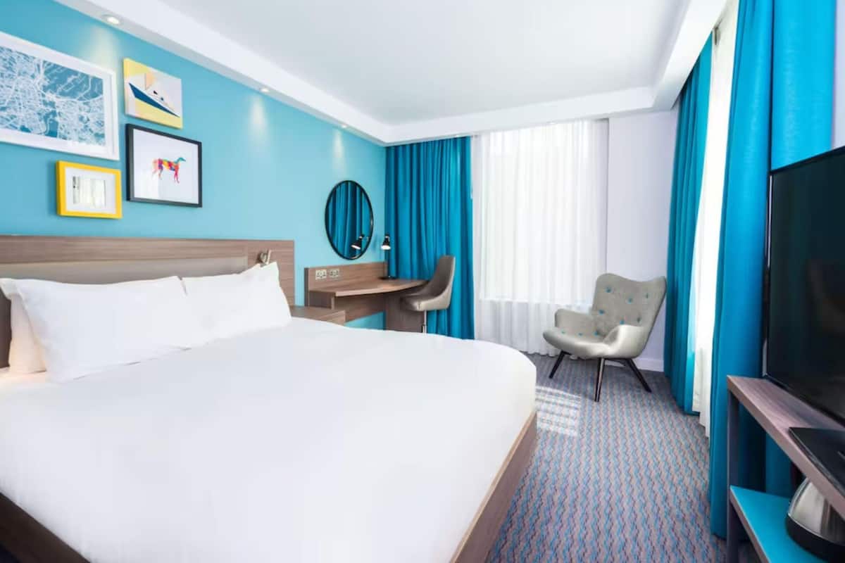 Une chambre d’hôtel avec un grand lit, un bureau, une chaise, une télévision et une fenêtre avec des rideaux.