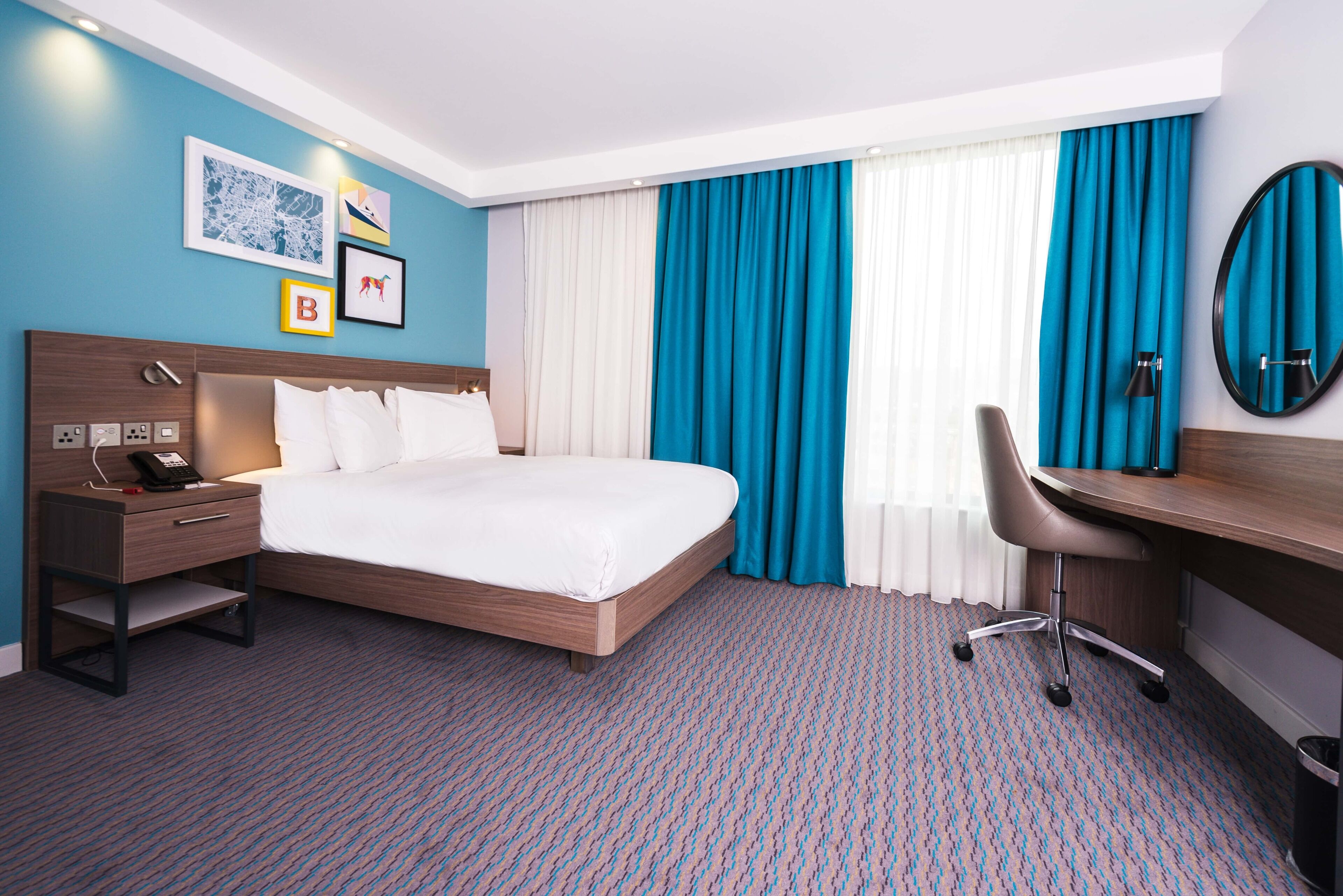 A hotel room with a bed, desk, chair, and a nightstand with a phone.