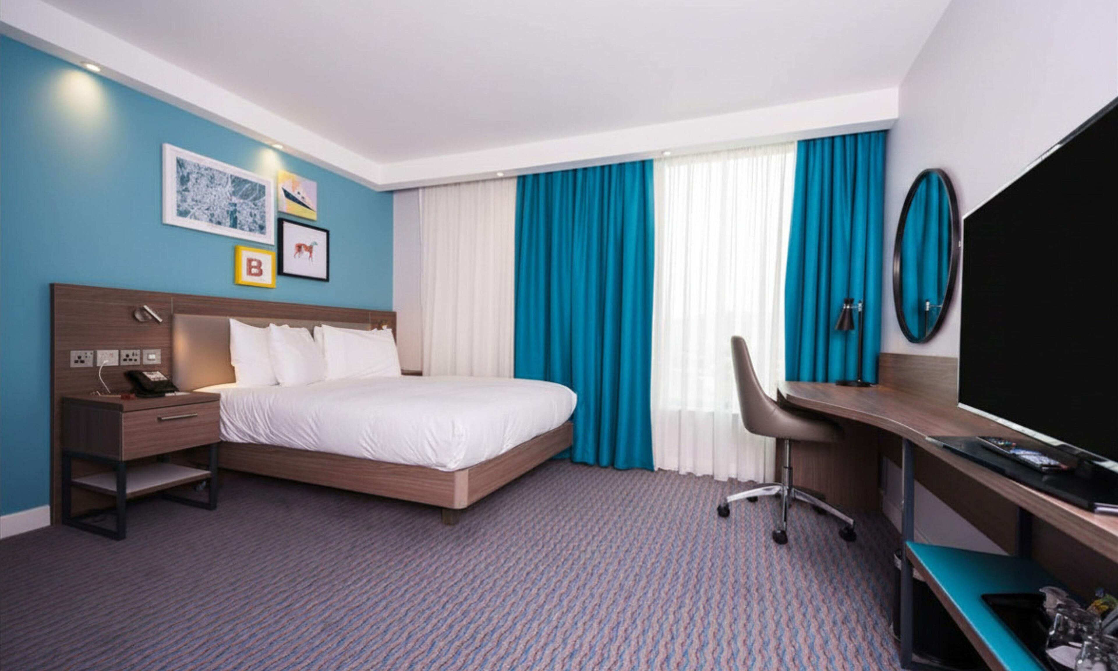 Una habitación de hotel con cama, escritorio, silla, televisor y una obra de arte en la pared.