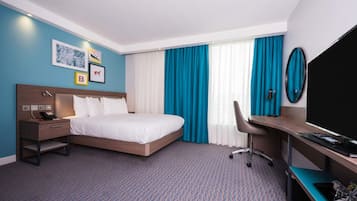 A hotel room with a bed, desk, chair, TV, and artwork on the wall.