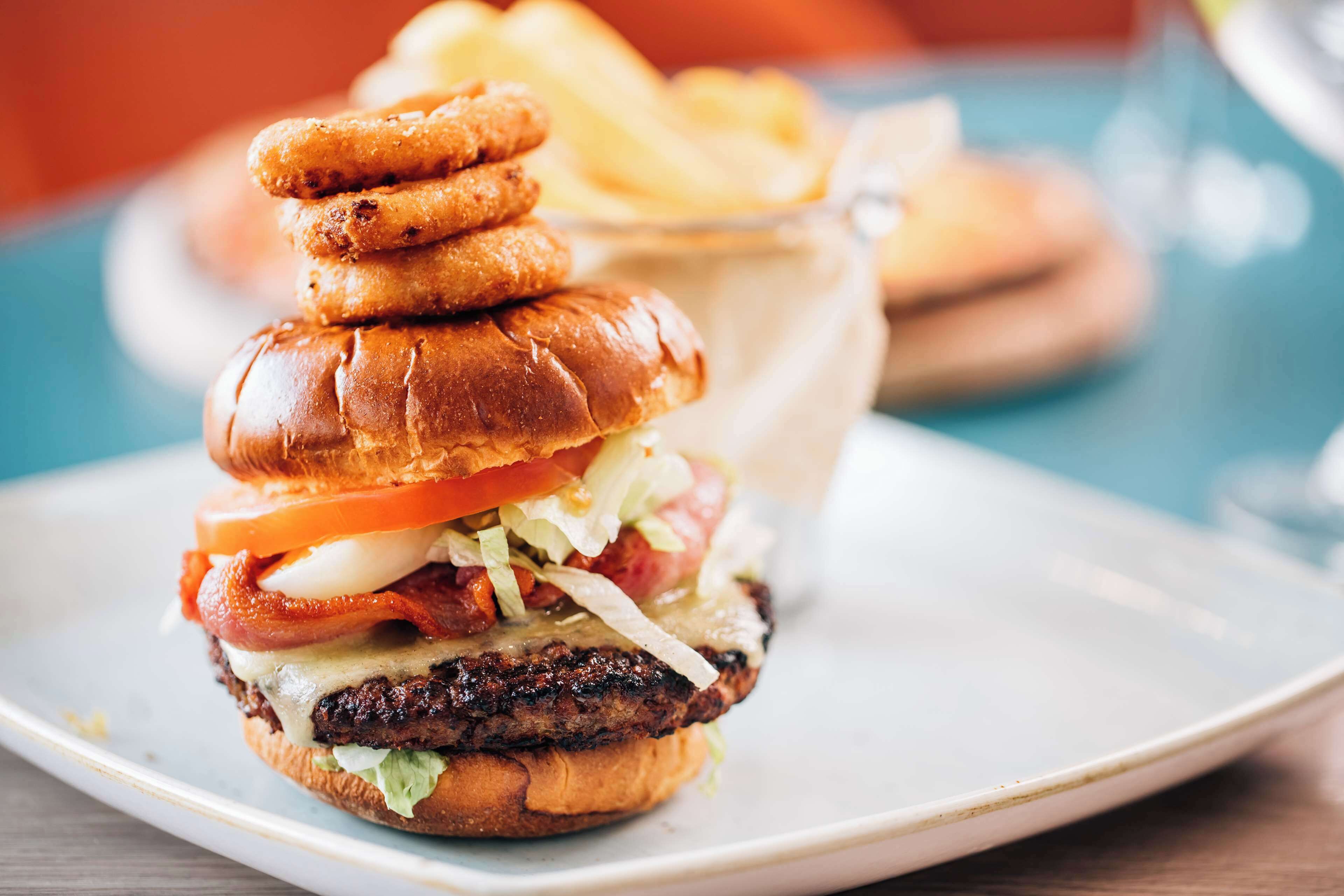 A burger with bacon, tomato, lettuce, and cheese on a bun, accompanied by fries and a drink in the background.