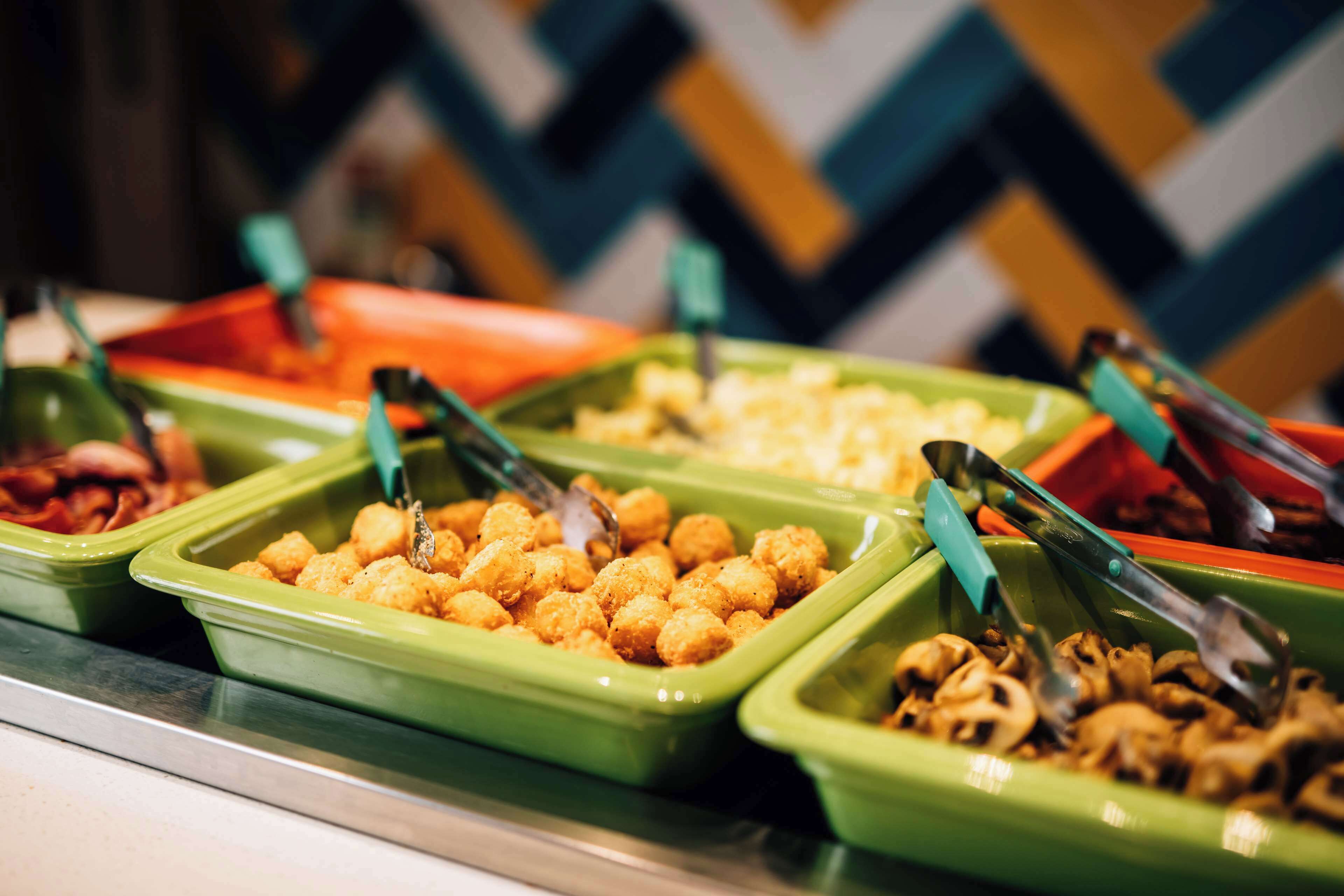Ein Buffet mit verschiedenen Speisen in grün und orangefarbenen Behältern, inklusive Küchenzangen zum Servieren.
