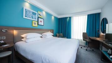 A hotel room with a large bed, a desk with a chair, a TV, and artwork on the wall.