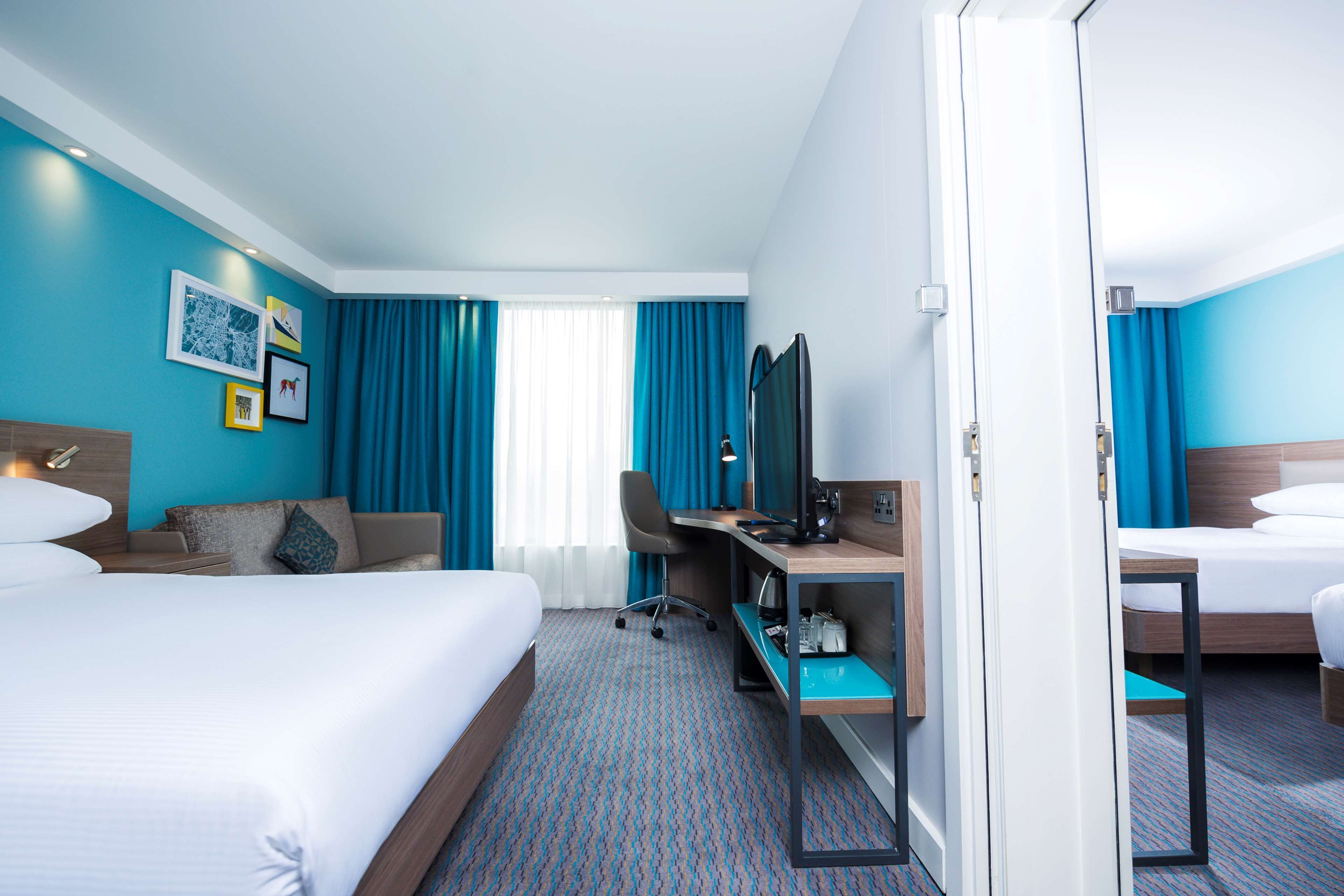 Ein Hotelzimmer mit zwei Betten, einem Schreibtisch, einem Sessel, einem Fernseher und einem Fenster mit Vorhängen.
