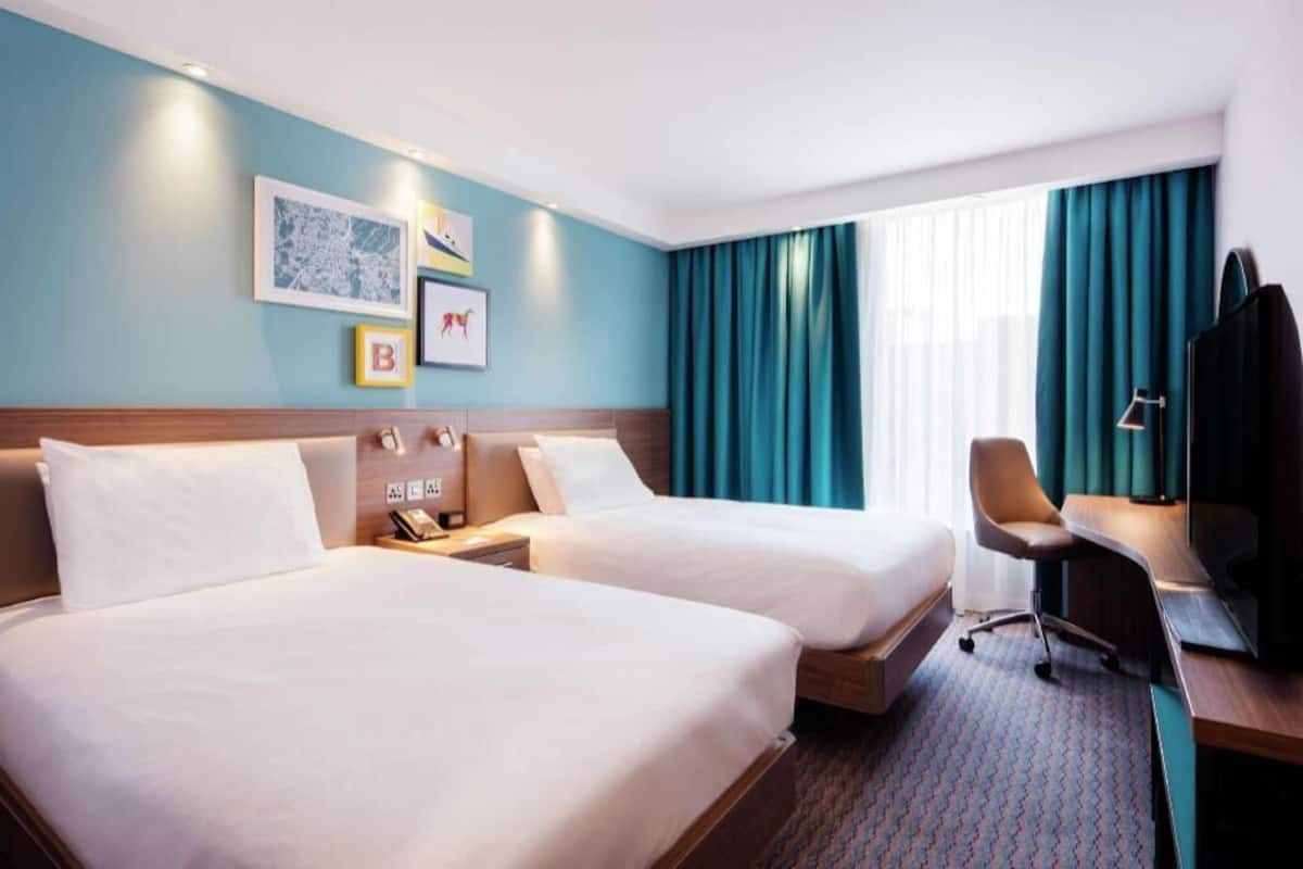 Une chambre d’hôtel avec deux lits, un bureau, une chaise, une télévision et des œuvres d’art accrochées au mur.