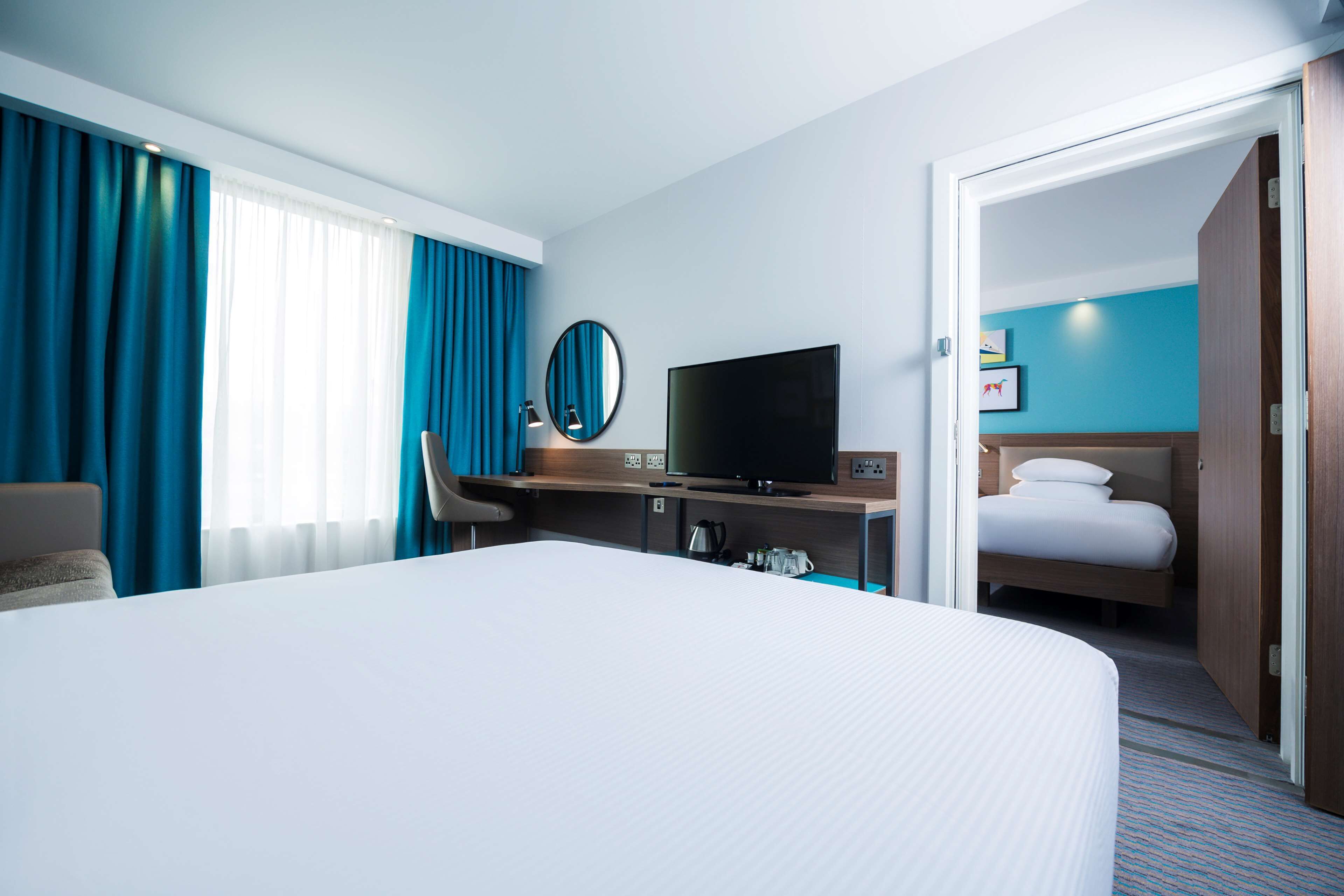 A hotel room with a large bed, a desk with a TV, a chair, and a door leading to another room.