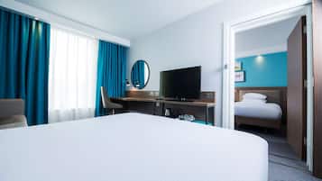 A hotel room with a large bed, a desk with a TV, a chair, and a door leading to another room.