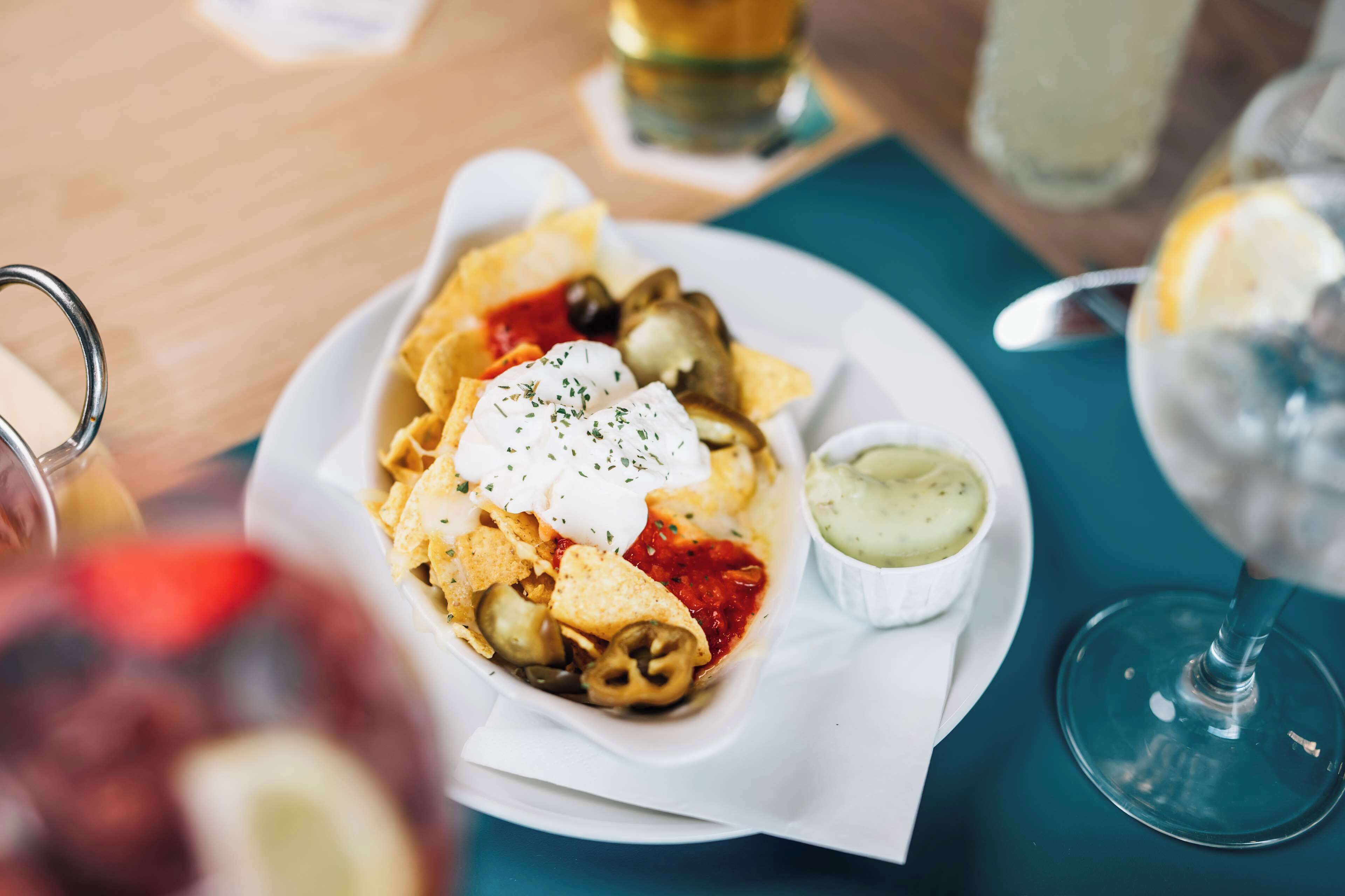 A plate of nachos with sour cream and jalapeños, a small bowl of sauce, and a glass of water on a table.