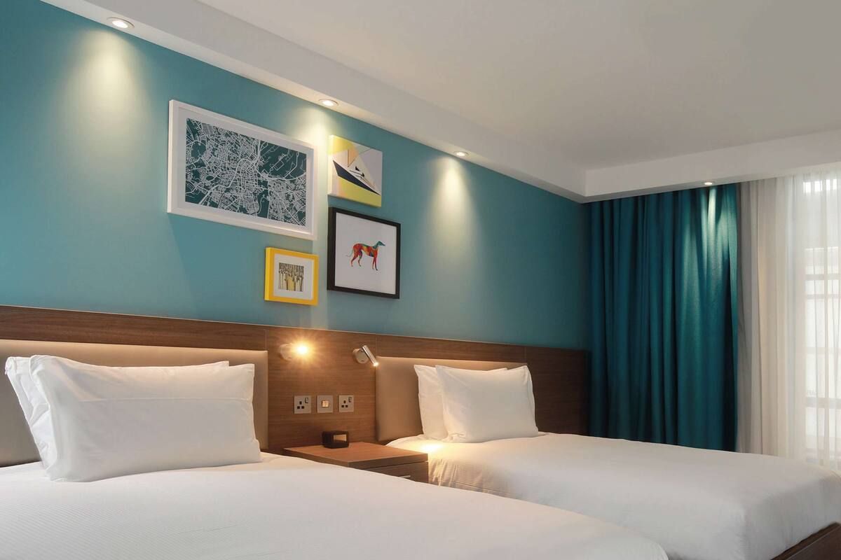 Une chambre d’hôtel avec deux lits, une tête de lit en bois, une table de chevet avec une lampe et des œuvres d’art accrochées au mur.