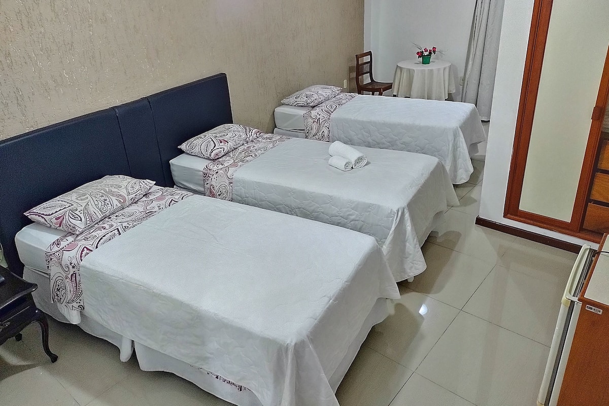 A hotel room with three beds, each with patterned bedding and a rolled towel on one. There is a small table with a vase of flowers and a chair.