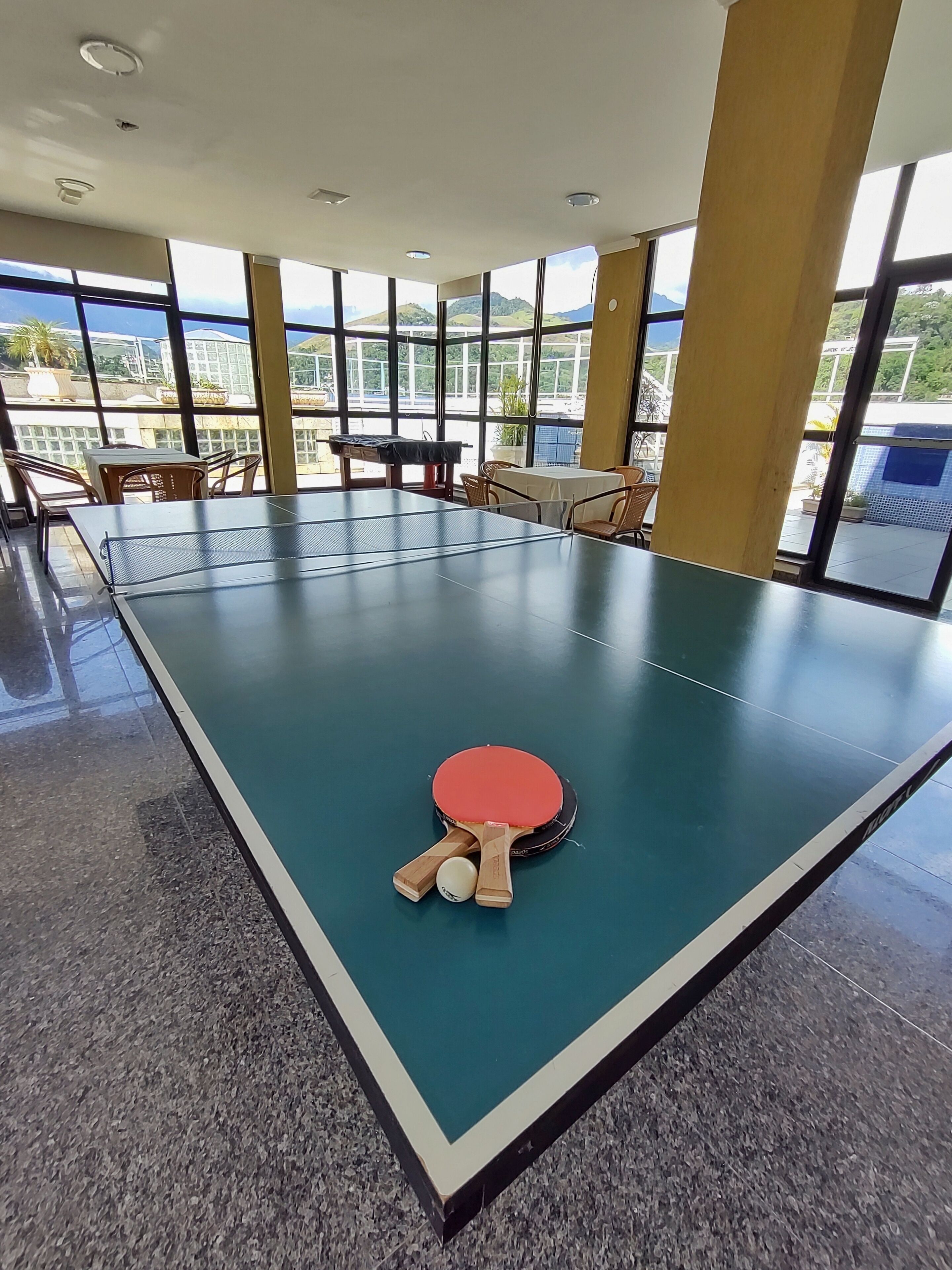 Un tavolo da ping pong con due racchette e una pallina in una stanza con ampie finestre e sedie.