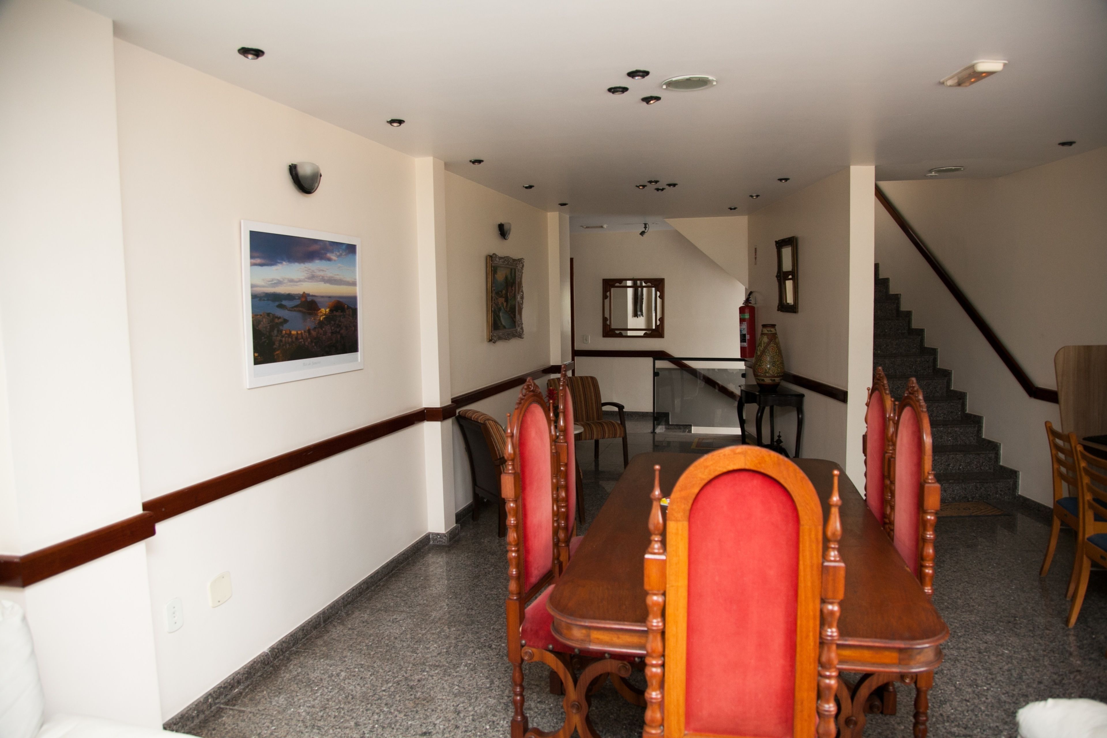 A dining area with a long wooden table and red upholstered chairs, a staircase in the background, and a framed picture on the wall.