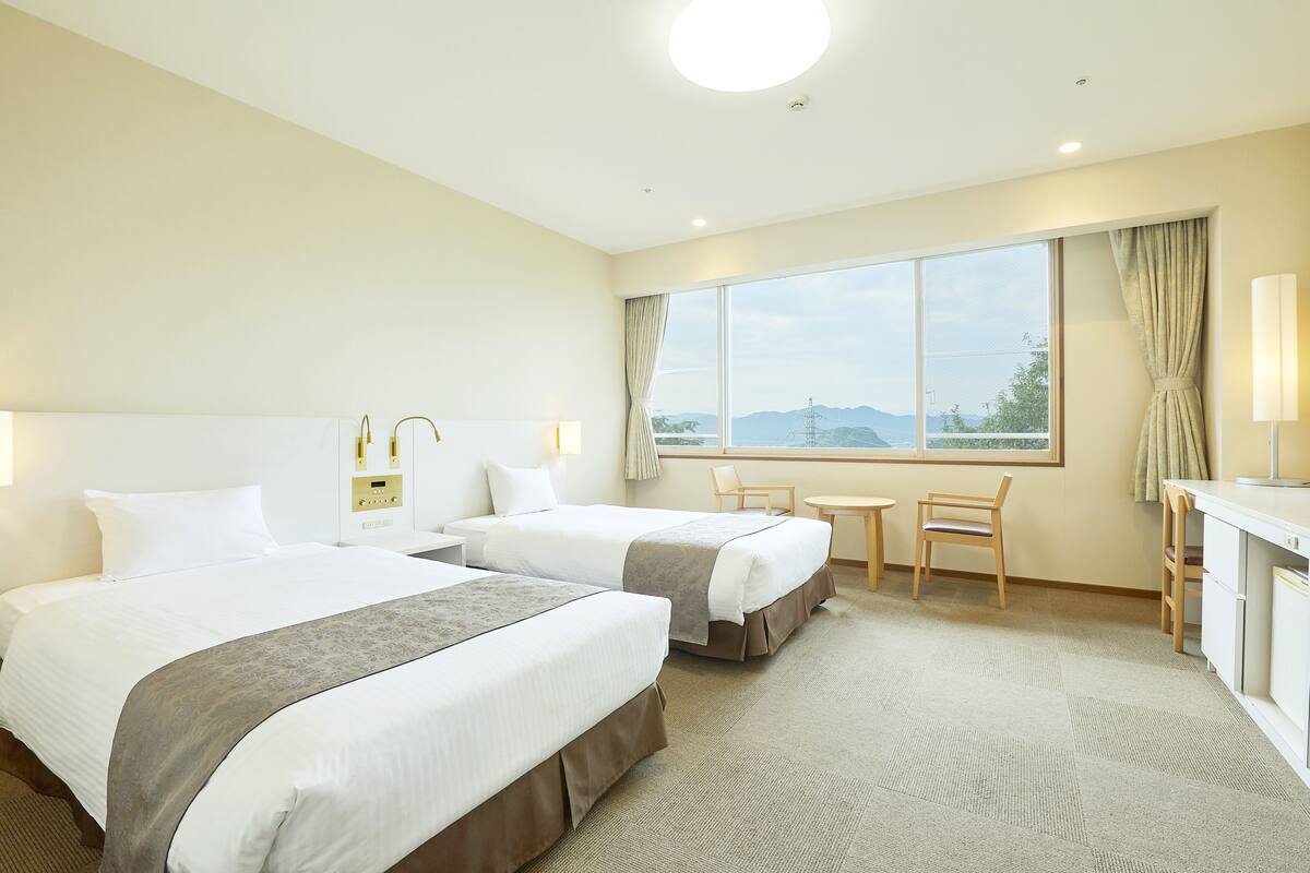 A hotel room with two beds, a desk, a chair, a small table, and a large window with a view.