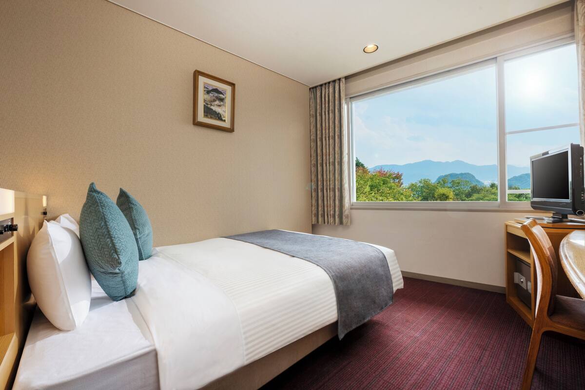 A hotel room with a bed, a desk with a computer, a window with a mountain view, and a framed picture on the wall.