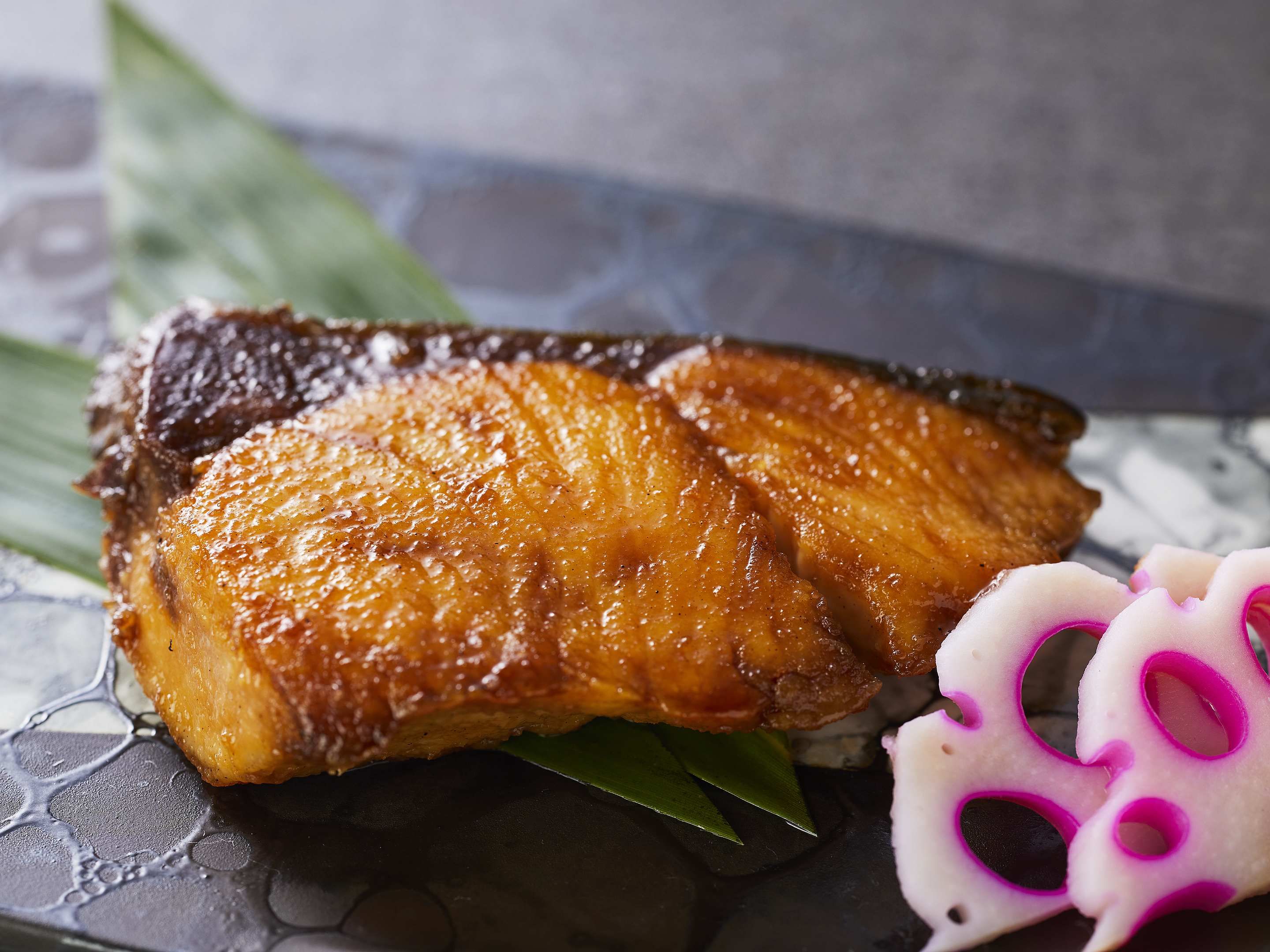 A close-up of a cooked pork belly with a glossy, caramelized surface, accompanied by a slice of white radish with purple patterns and a green leaf.