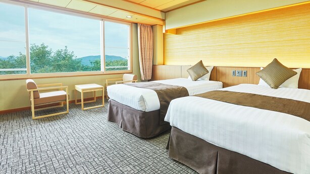 Habitación de hotel con dos camas, una mesita, una silla y un ventanal con vistas a zonas verdes.