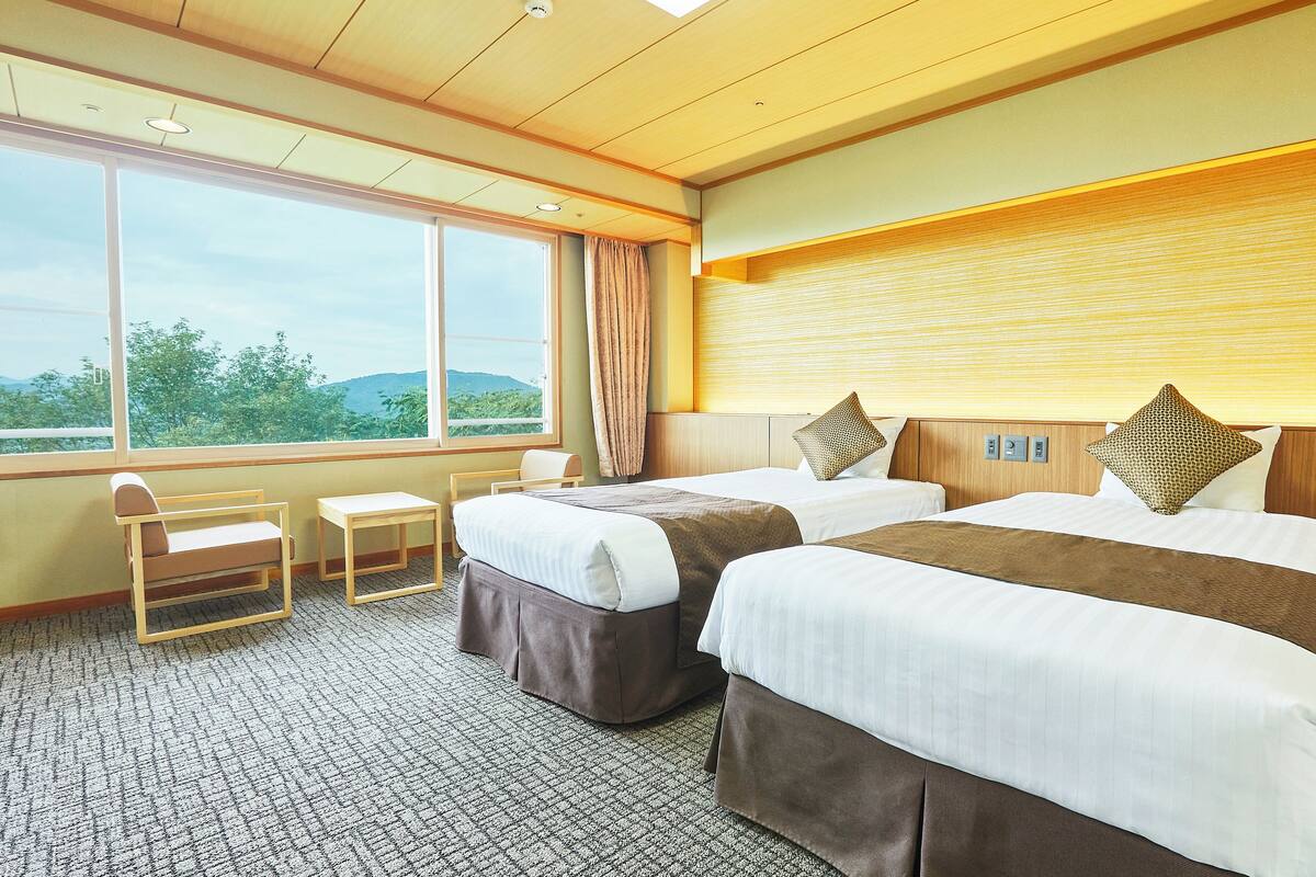 A hotel room with two beds, a small table, a chair, and a large window with a view of greenery.