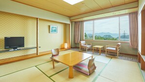 Ein traditioneller japanischer Raum mit Tatami-Boden, einem niedrigen Tisch, Stühlen, einem Fernseher und einem großen Fenster mit Blick auf Berge.