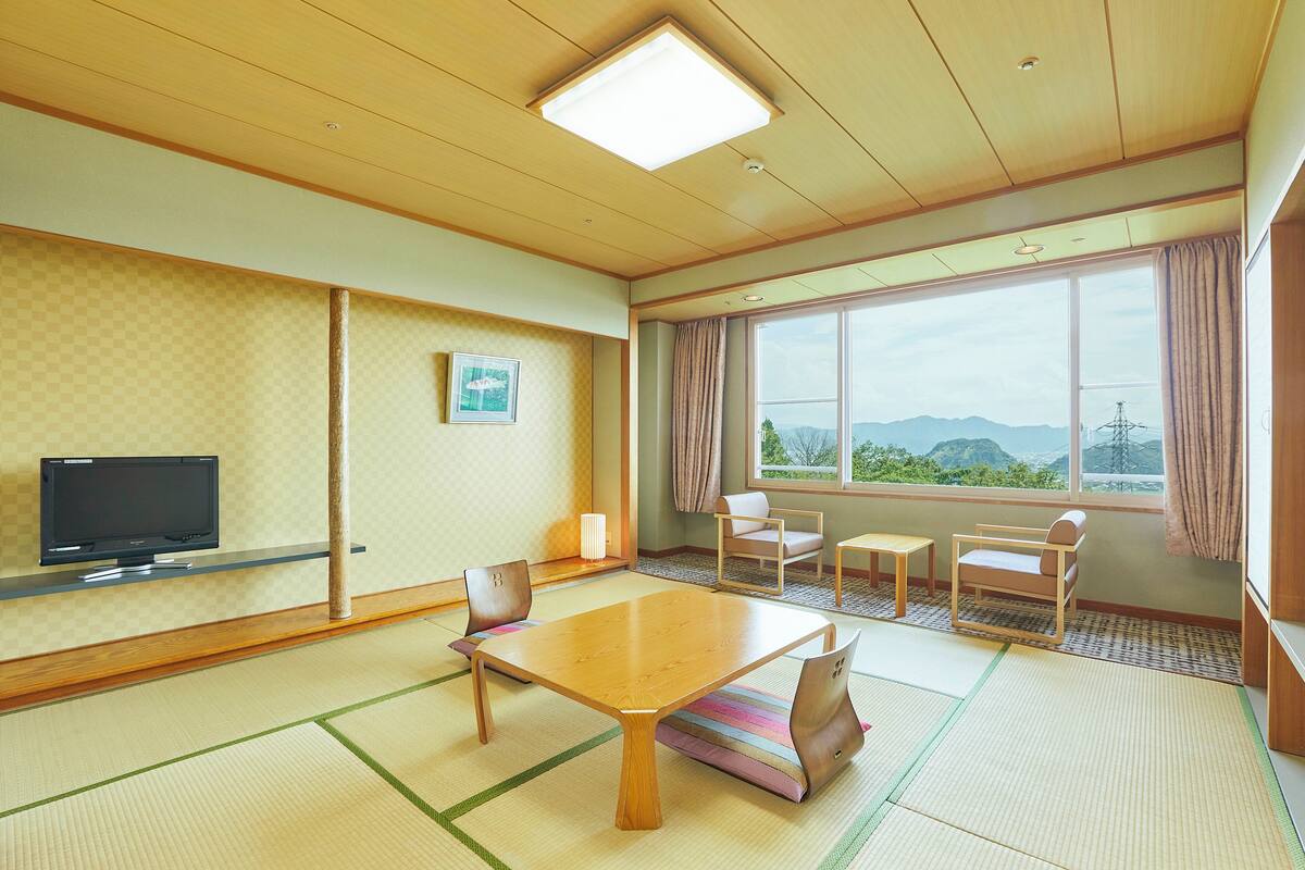 A traditional Japanese room with tatami flooring, a low table, chairs, a television, and a large window with a view of mountains.