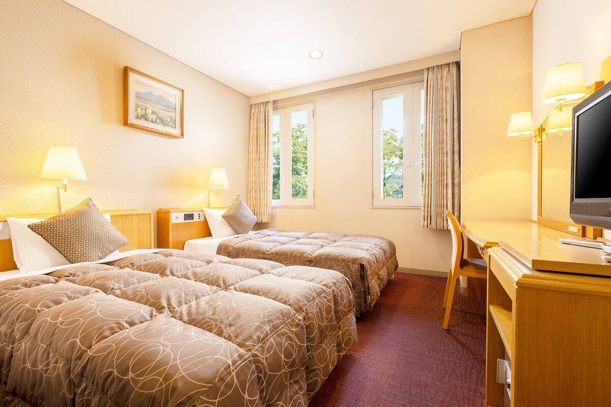 A hotel room with two beds, a desk, a television, and two windows with curtains.