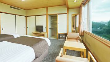 A hotel room with two beds, a TV, a small table, and a window with a view of greenery.