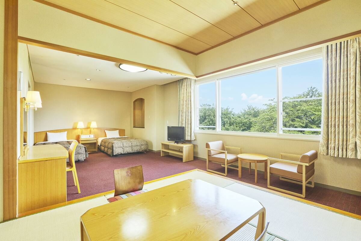 A hotel room with a bed, desk, chairs, a TV, and a large window with a view of trees.