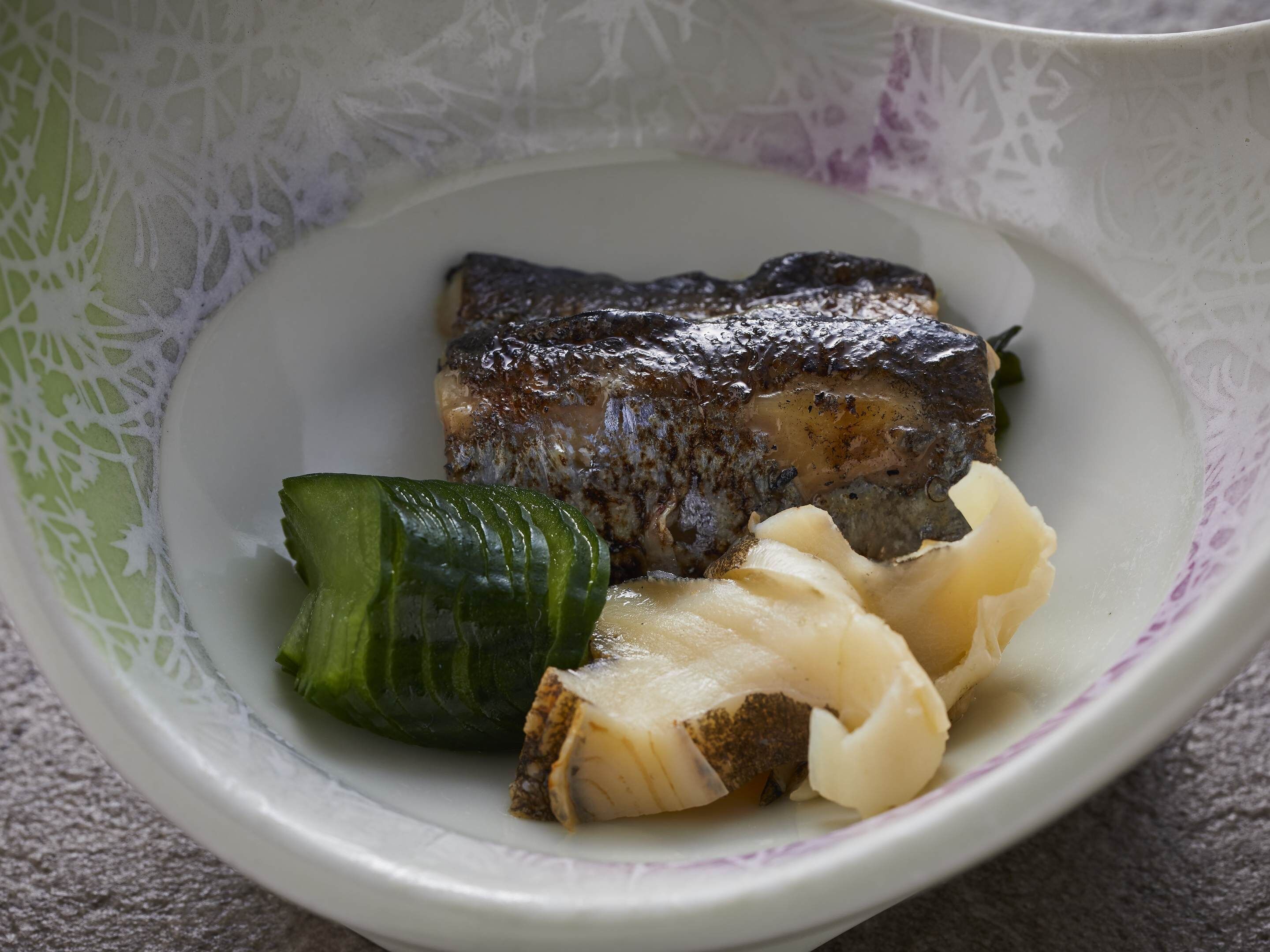 Un plato con un trozo de pescado a la parrilla, una rodaja de pepino y un trozo de pescado blanco.