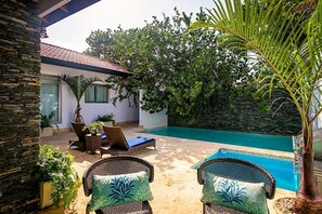 A poolside area with wicker chairs and a lounge chair, surrounded by greenery and a stone wall. - Villa Caliche by Unlimited Luxury Villas (La Romana)