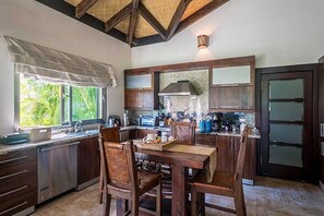 A kitchen with wooden cabinets, a stainless steel dishwasher, a dining table with chairs, and a window with a view of greenery. - Villa Caliche by Unlimited Luxury Villas (La Romana)