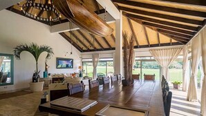 Un vaste espace dédié à la salle à manger, avec une longue table en bois, des chaises, un plafond doté de poutres apparentes et un luminaire suspendu.