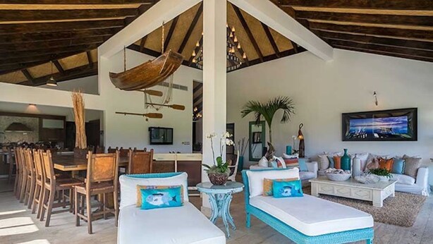 Un vaste espace de vie doté d’un plafond haut, de poutres en bois, d’un coin repas avec des chaises en bois, d’un ensemble de canapés blancs et d’une décoration en forme de bateau suspendu.