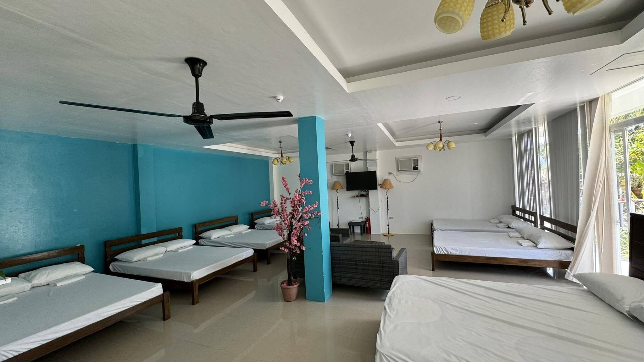Habitación de hotel con cuatro camas individuales, ventilador de techo, televisión y un amplio ventanal con cortinas.