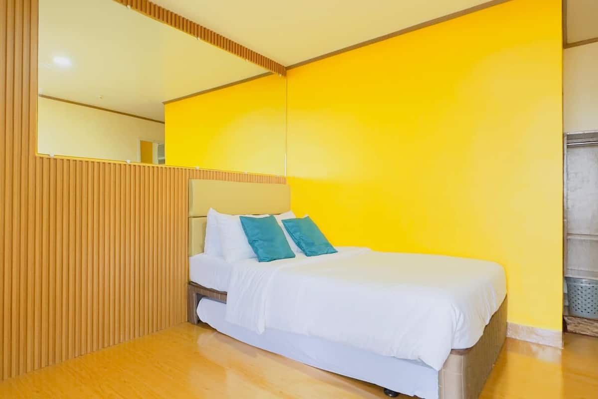 A modern hotel room with a wooden floor, a bed with white bedding and teal pillows, and a bright yellow accent wall.