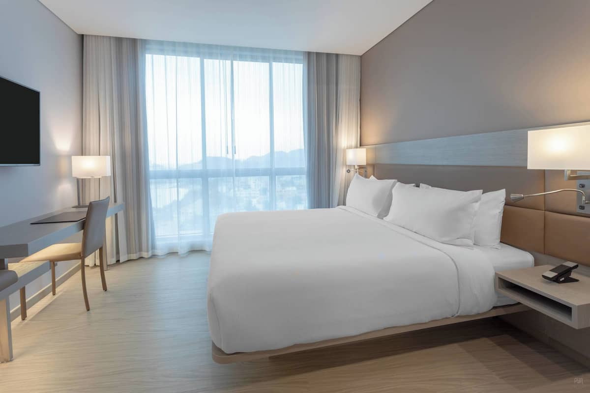 Habitación de hotel moderna con una cama grande, un escritorio y una silla. Hay un televisor montado en la pared y una ventana con cortinas.