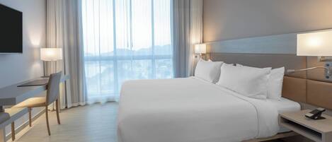 Habitación de hotel moderna con una cama grande, un escritorio y una silla. Hay un televisor montado en la pared y una ventana con cortinas.